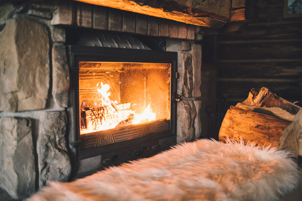 A cozy fireplace with a burning fire is seen inside a stone and wood-paneled room, with a fur-covered seat in the foreground.